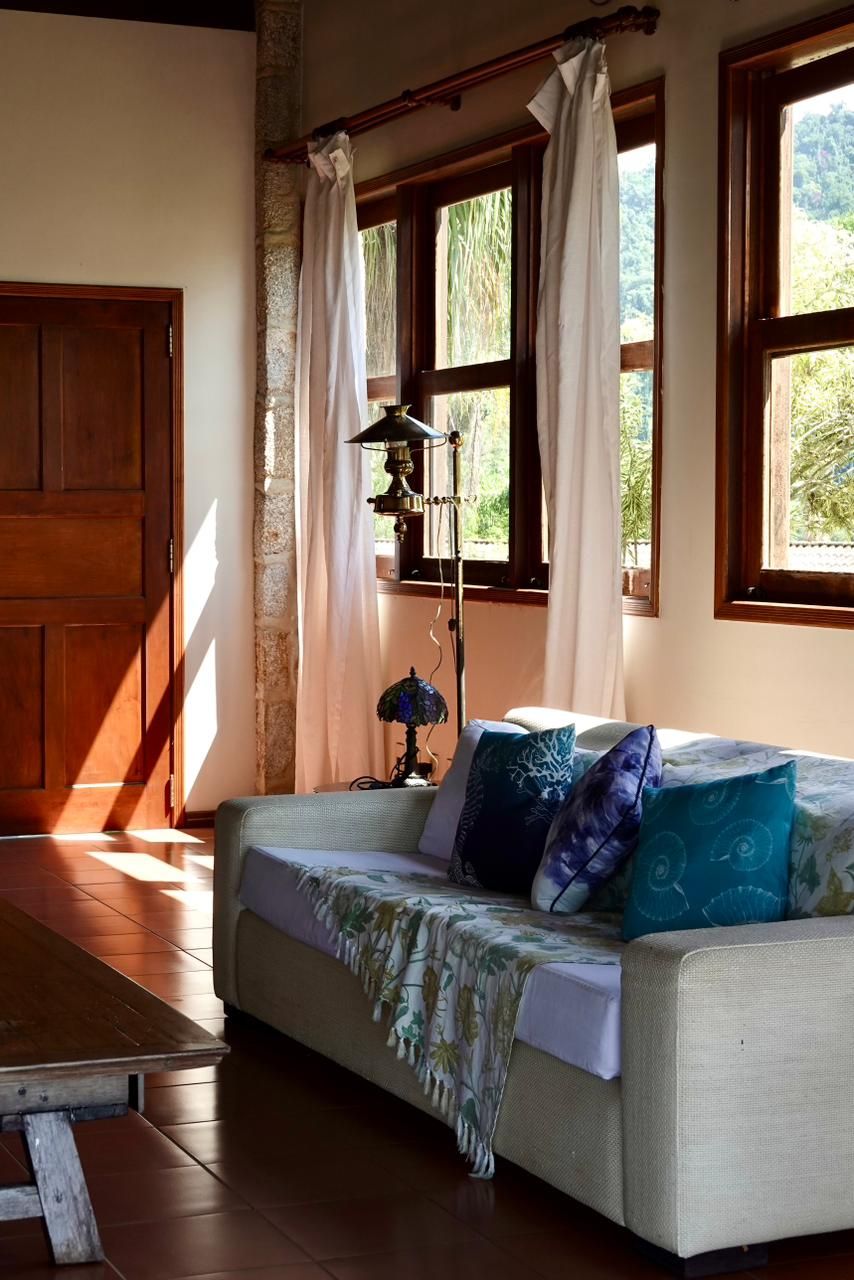 Sala de estar ensolarada com sofá creme, almofadas azuis, janelas amplas e piso de madeira.