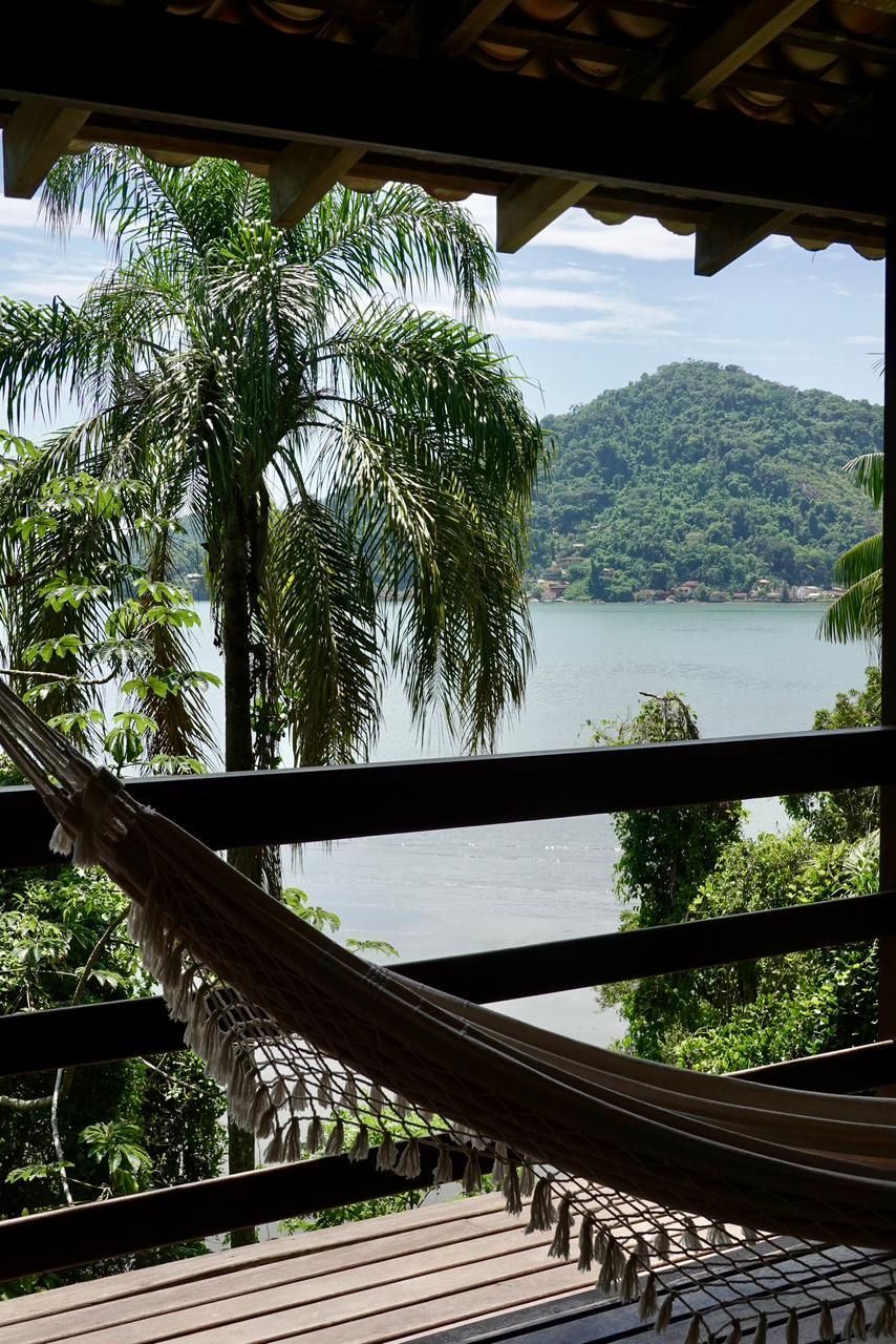 Rede em uma varanda coberta com vista para um lago tranquilo e uma encosta verdejante através de grades abertas.