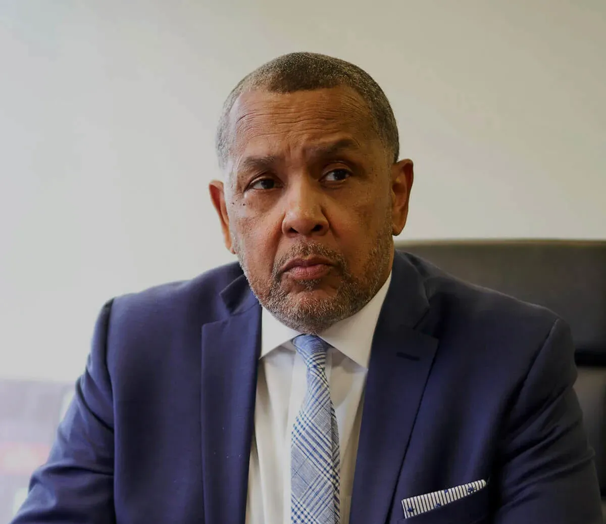 A man in a blue suit, white shirt, and patterned blue tie, looking slightly to the side against a neutral background.