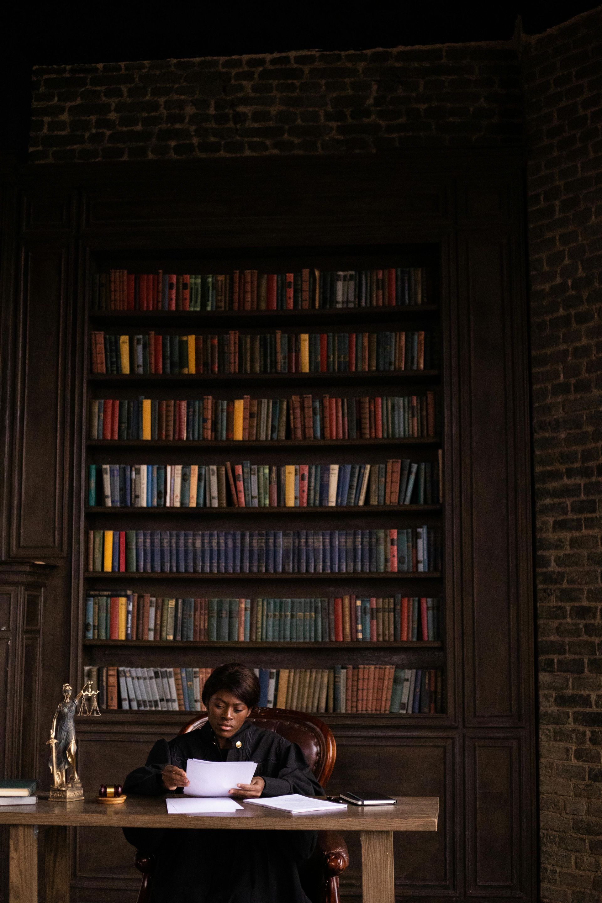 A person in a black judicial robe sits at a wooden desk reading documents in a library with tall shelves of books.