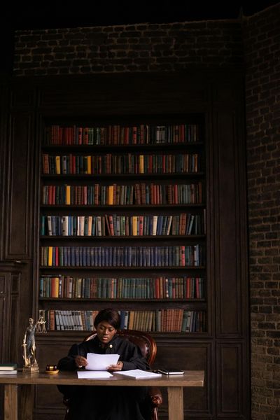 A person in a black judicial robe sits at a wooden desk reading documents in a library with tall shelves of books.