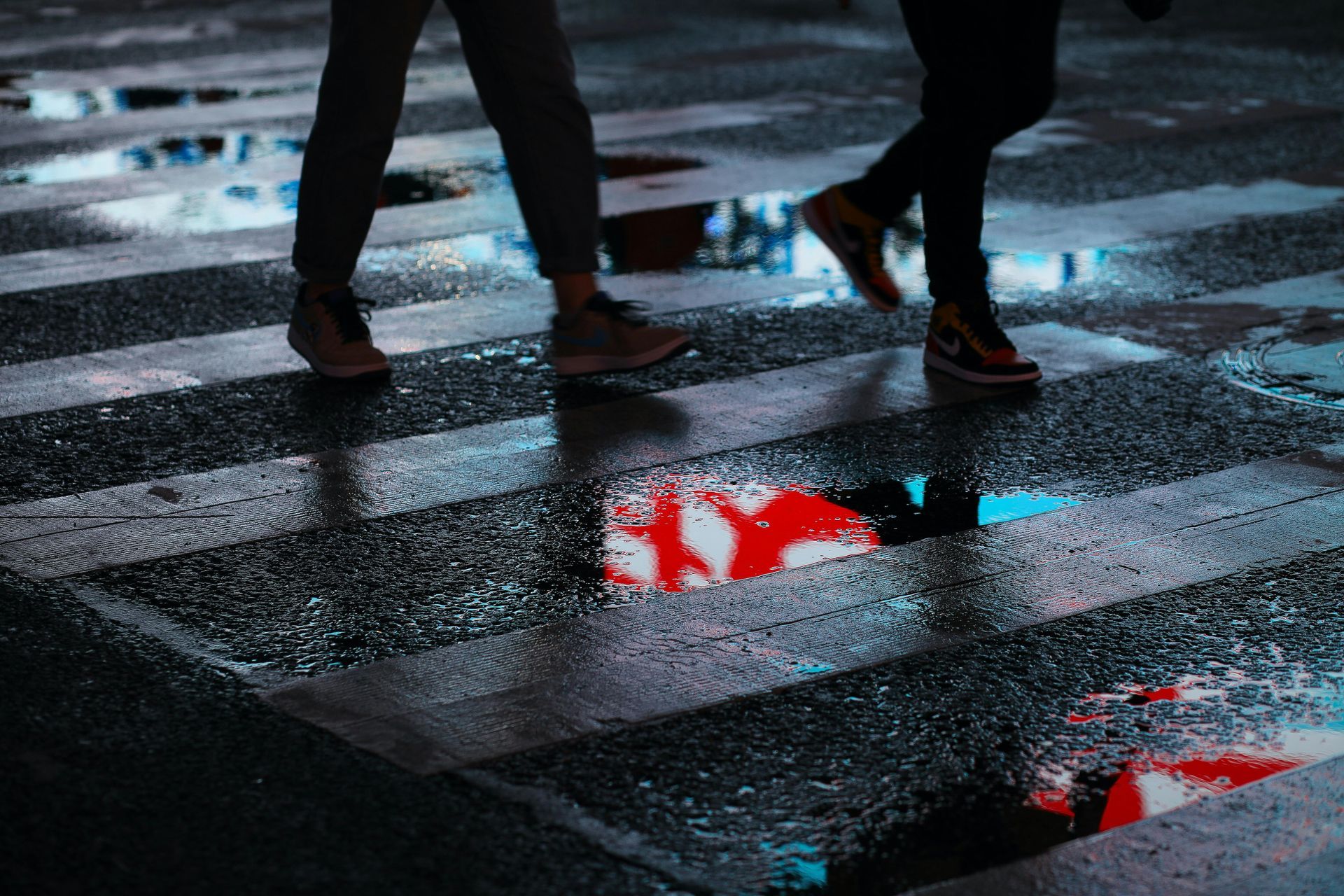 Two people walk across a wet, rain-slicked crosswalk, their feet reflected in puddles showing bright red traffic signals.