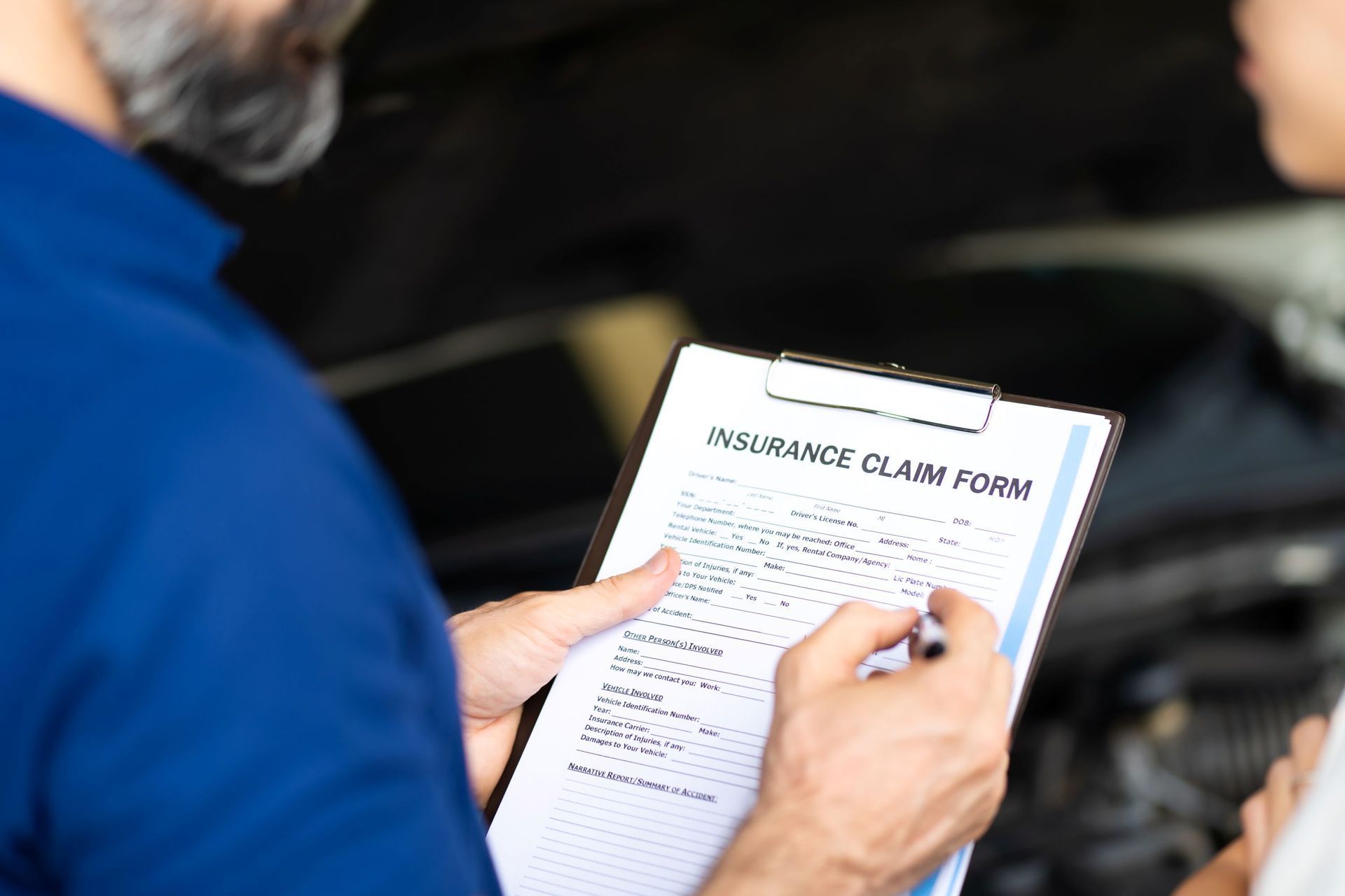 A person in a blue shirt holds a clipboard with an insurance claim form while standing in front of a car engine.