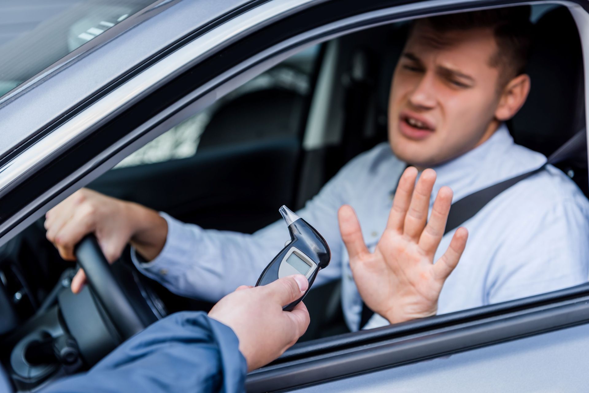 A driver in a car holds up a hand to refuse a breathalyzer test being presented by an officer.