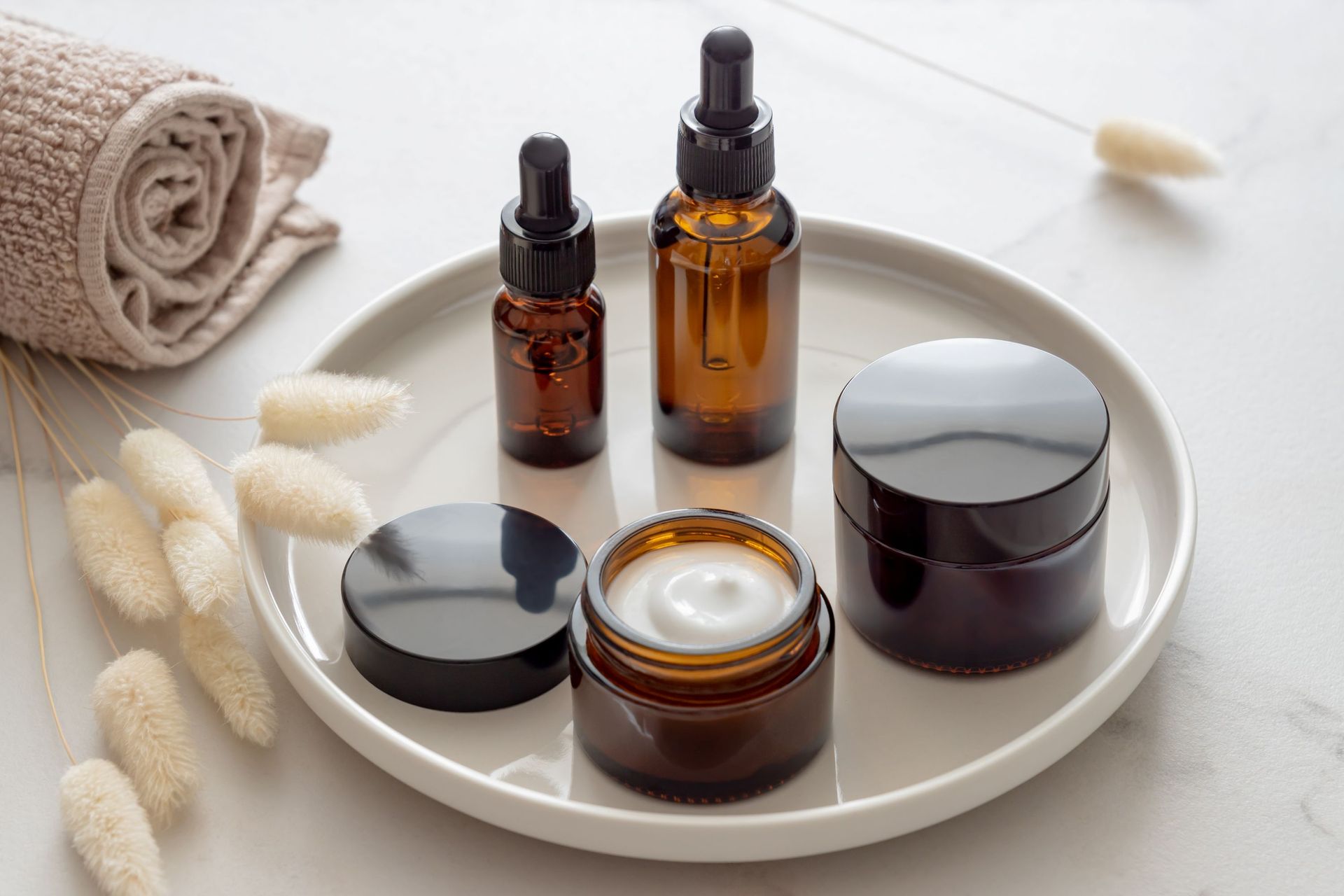 Amber glass serum bottles and jars of cream on a white plate, alongside a rolled towel and dried botanical stems.