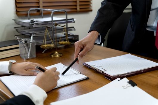 A person in a suit points to a document while another person prepares to write at a wooden desk with law scales nearby.