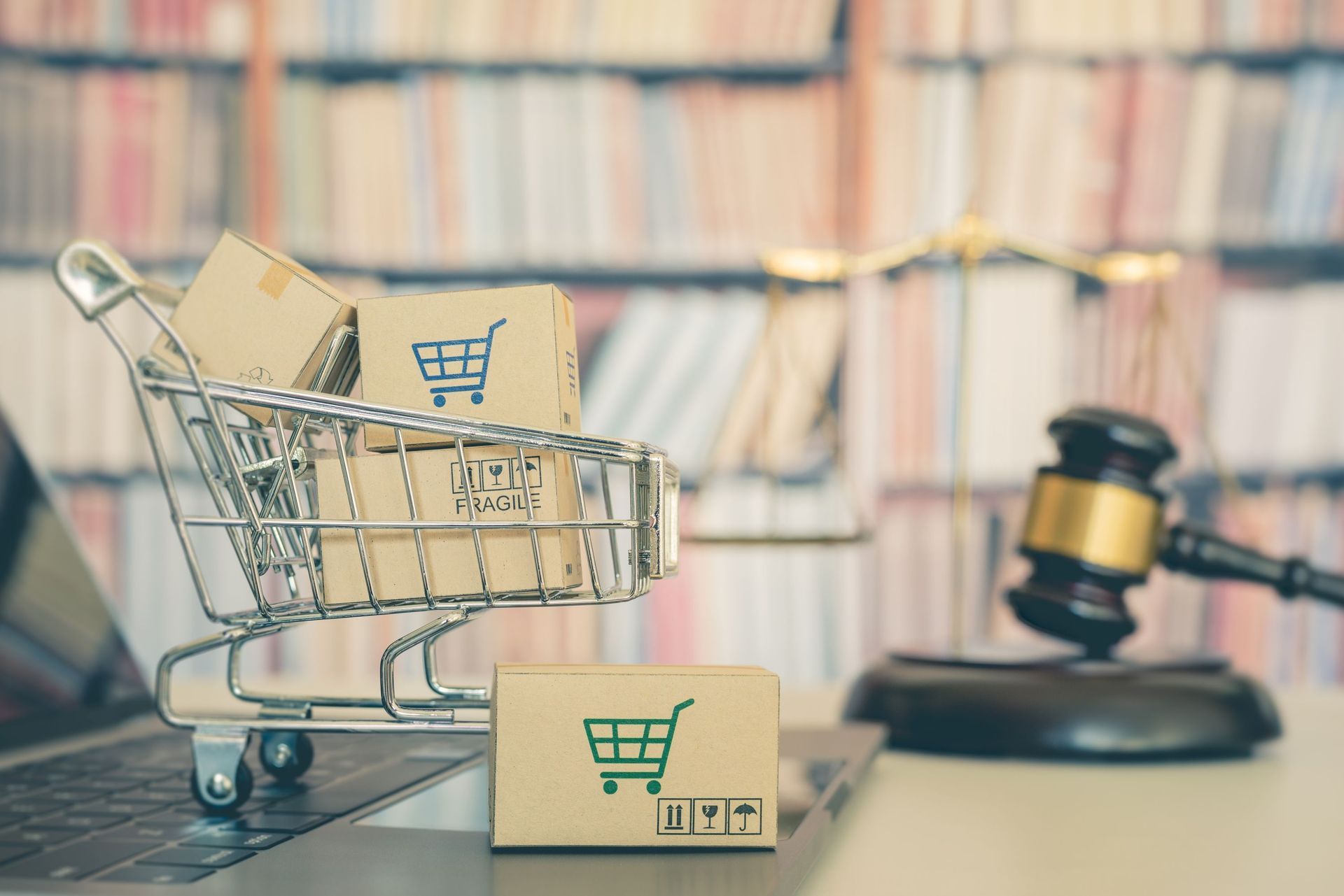 A small shopping cart with cardboard boxes on a laptop, with a judge's gavel and a blurry library background.
