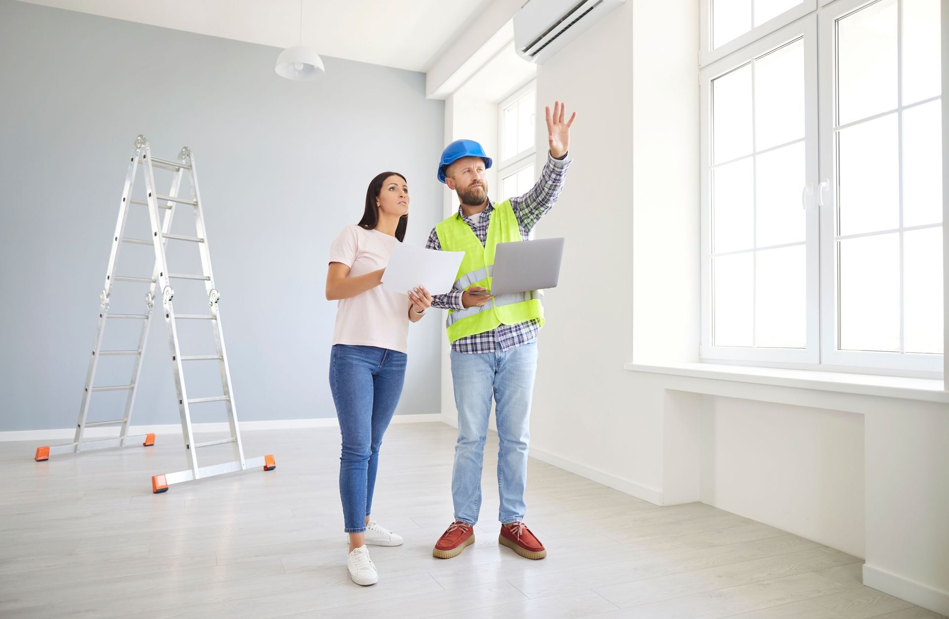 A construction professional in a hard hat and vest points at a window while discussing renovation plans with a colleague.