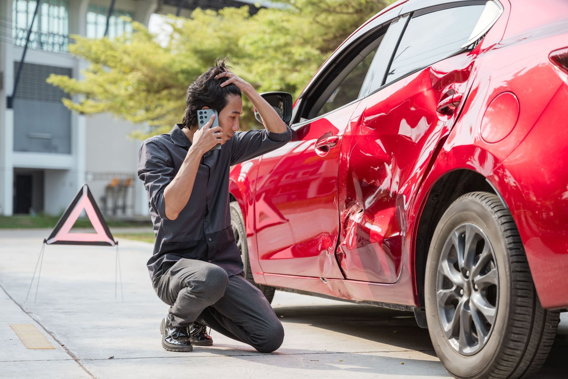 A person kneels next to a red car with significant side-door damage, looking distressed while talking on a phone.