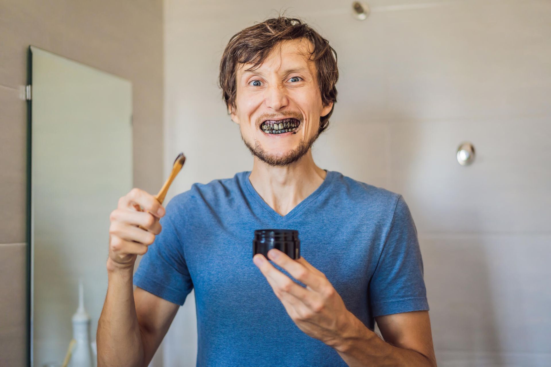 Smiling man in a blue shirt brushing his teeth and holding a toothpaste tube in a bathroom mirror.