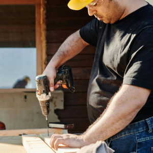 Man using a drill to work on a wooden board outside a cabin