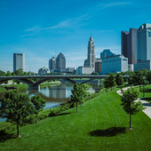 Riverfront park with a bridge, green lawn, trees, and city skyline under a blue sky