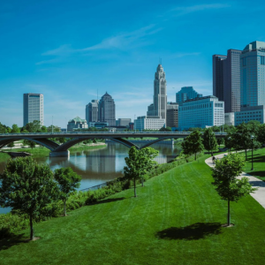 Riverfront park with a bridge, green lawn, trees, and city skyline under a blue sky