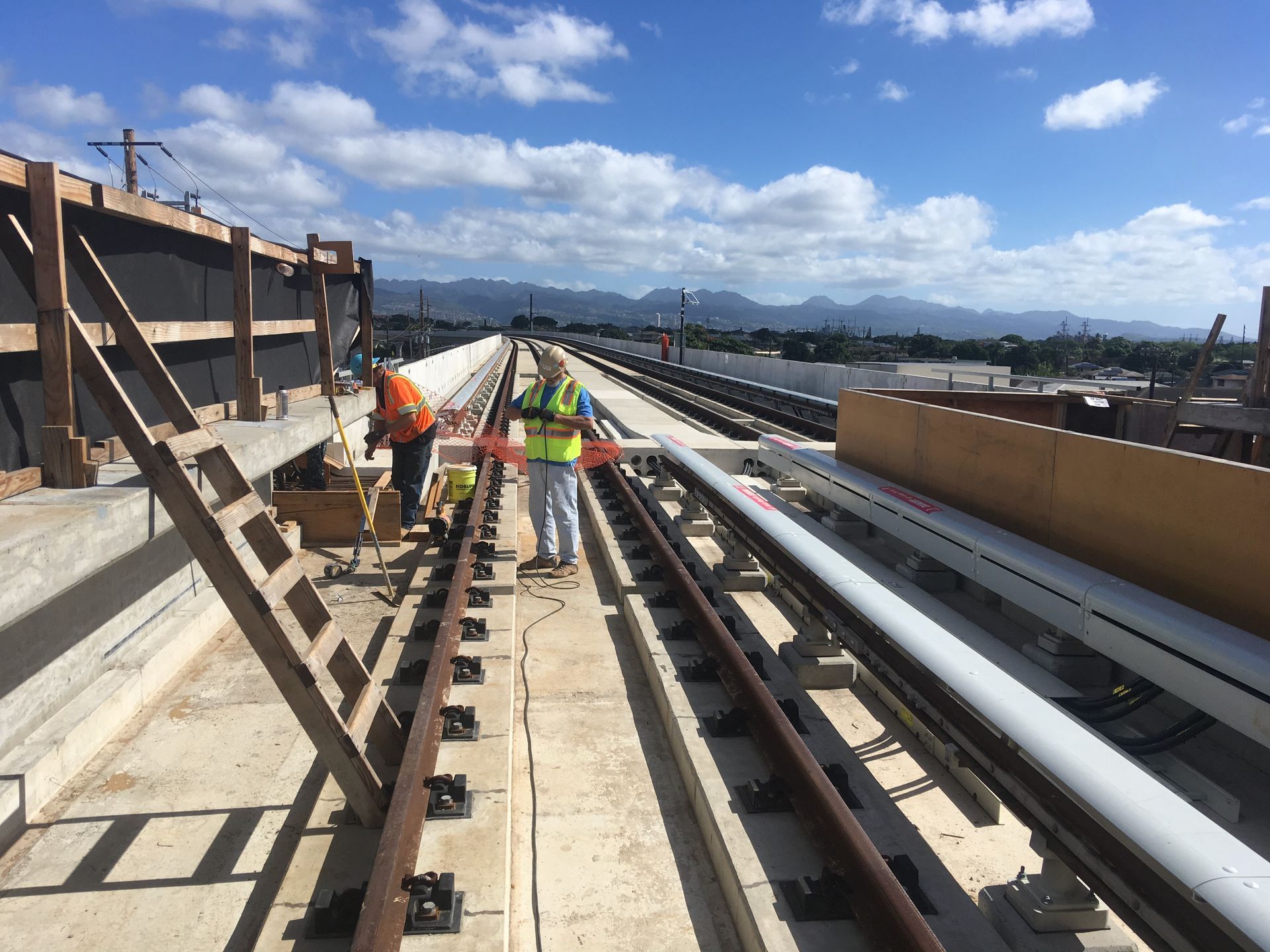 Construction workers beside elevated rail tracks at Pouhala station, with Oʻahu mountains in the distance.