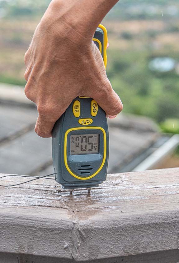 A handheld moisture meter probes the painted timber railing.