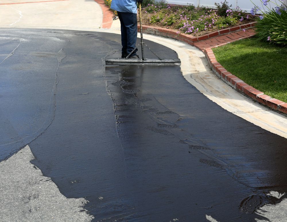 A person is spreading asphalt on a driveway with a broom.
