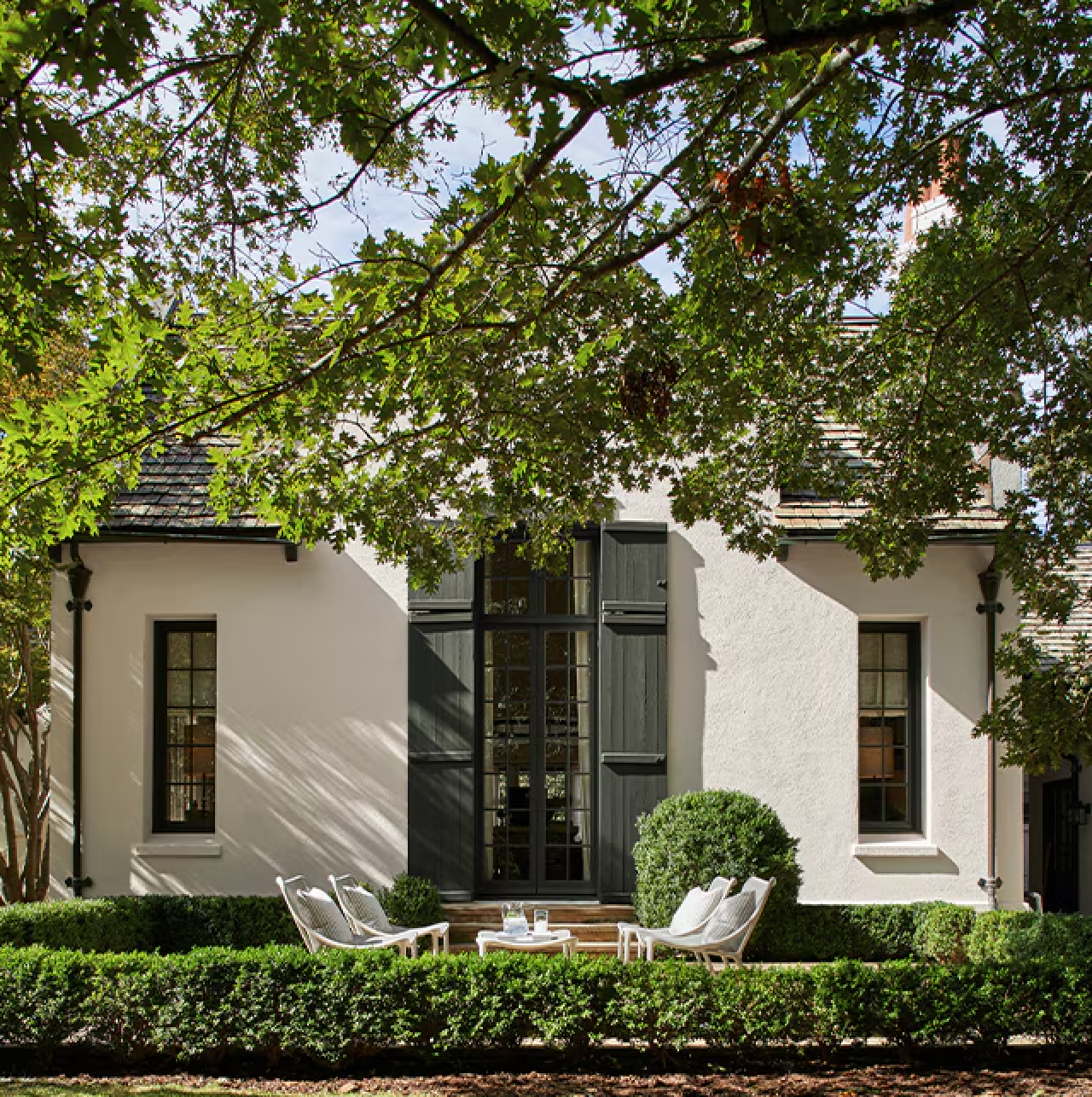 White house facade with black shutters, lush hedges, and a shaded front patio under trees