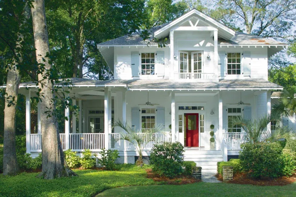 White two-story house with a red front door, white porch, and large trees in a green yard