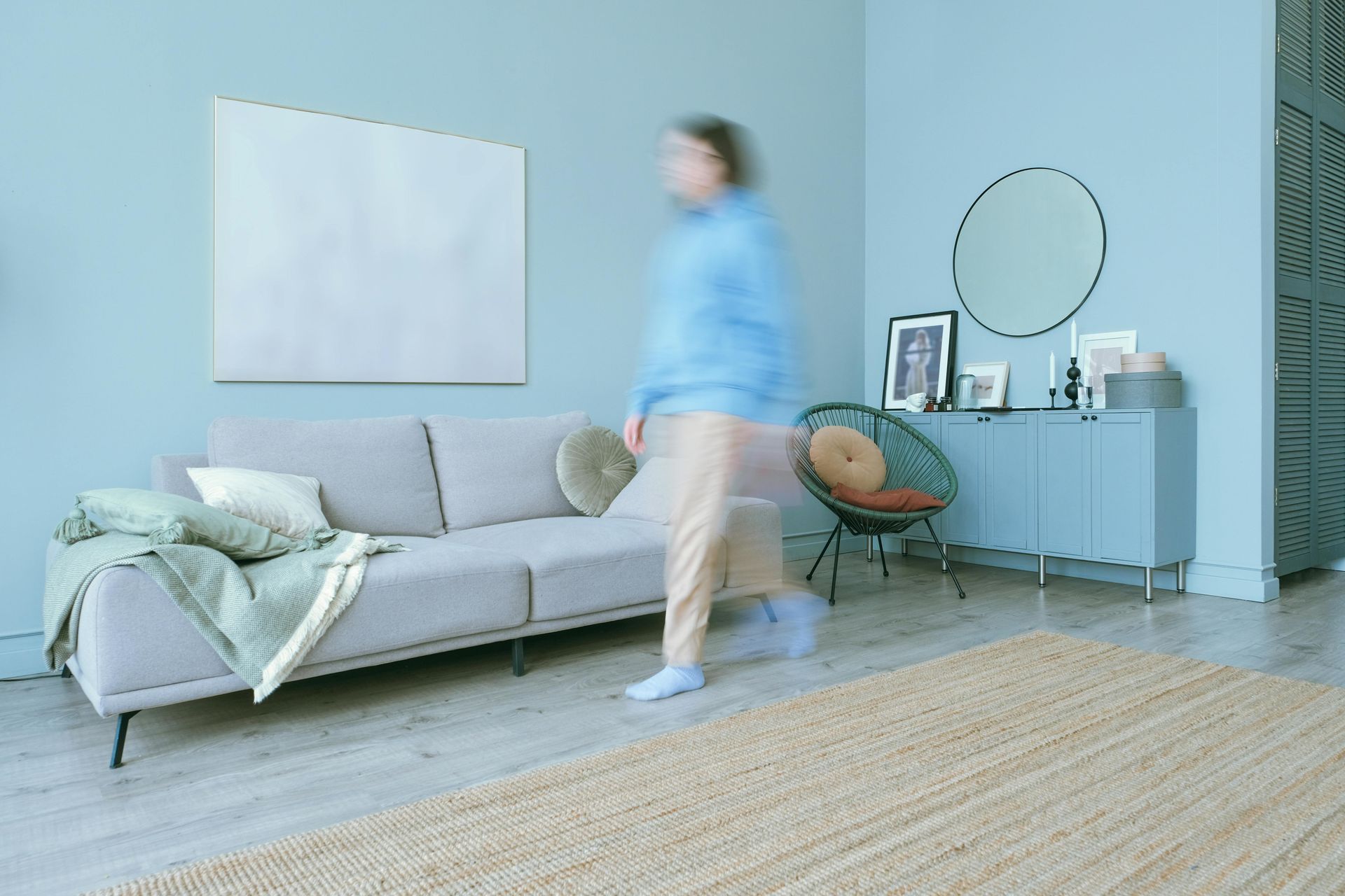 Blurred person walks in a light blue room with a sofa, rug, and cabinet.