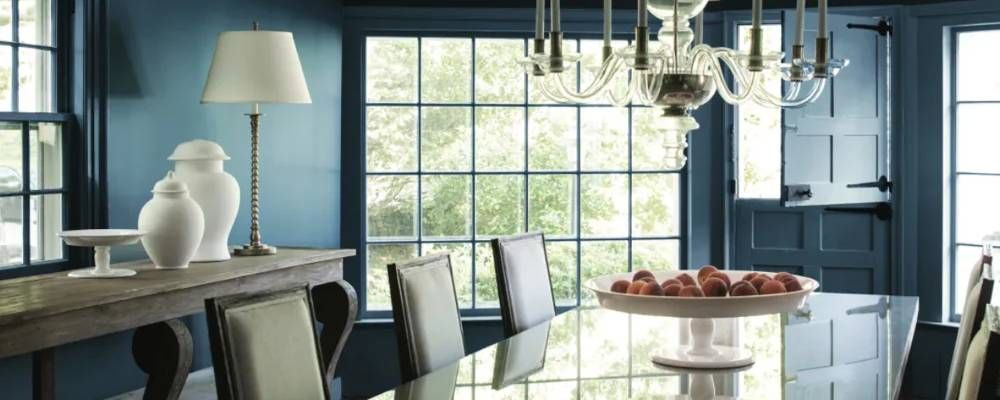 Elegant dining room with blue walls, glass table, chandelier, and vase centerpiece near a window.