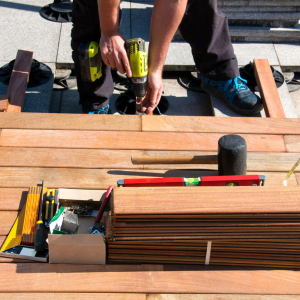Person using a power drill on wooden framing at a rooftop worksite, with tools and lumber stacked nearby
