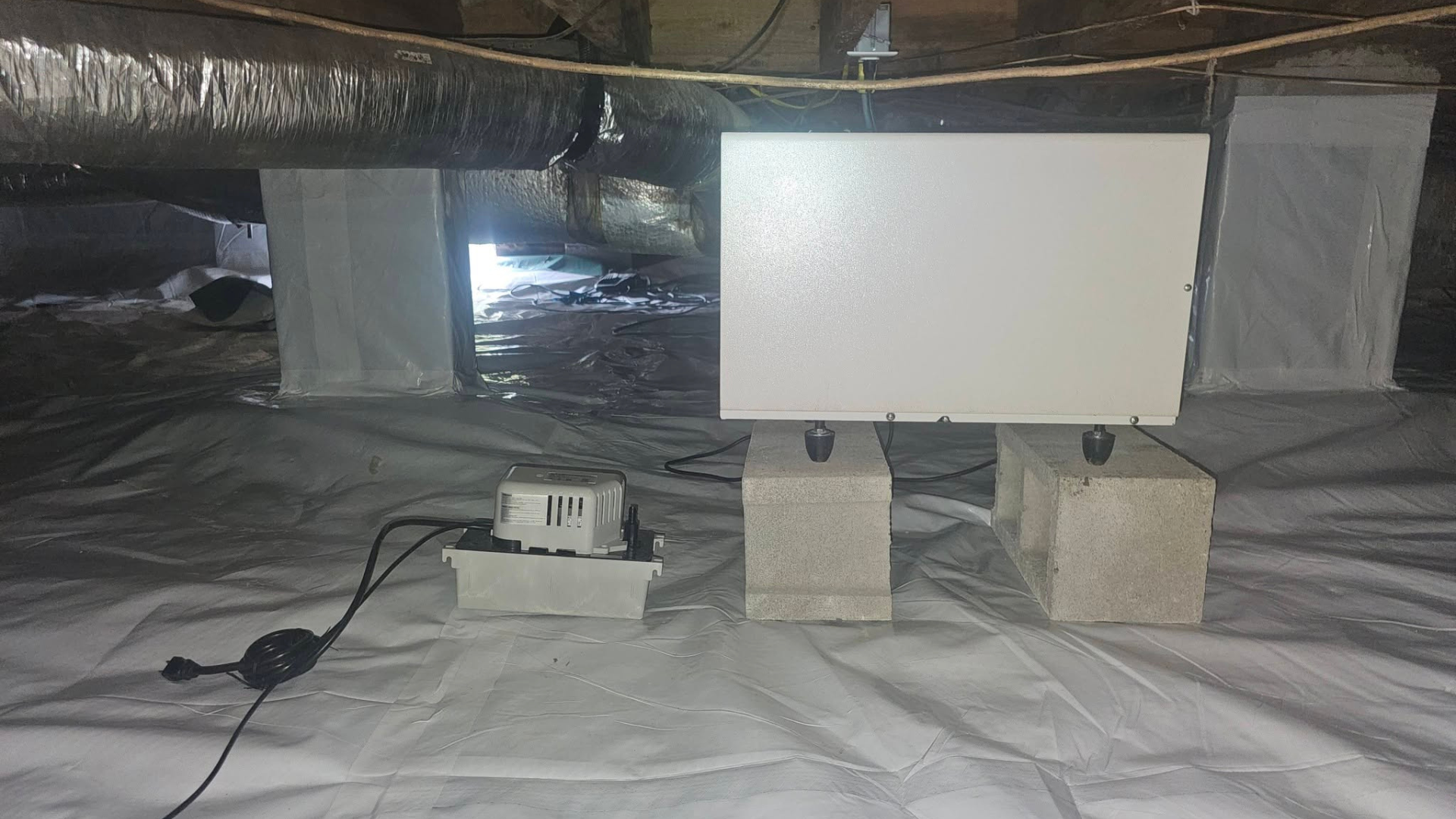 Large white rectangular panel on blocks in an attic, with a small device and cables on a plastic-covered floor.