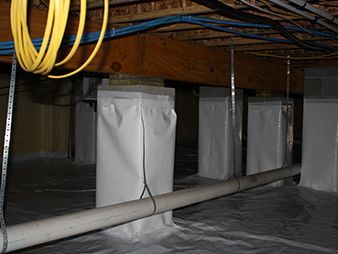 Crawlspace under a house with wrapped support posts, exposed pipes, and hanging cables