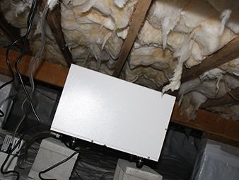 White electrical panel mounted among exposed insulation, pipes, and wires in an attic or crawl space.