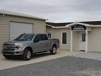 Silver pickup truck parked outside a beige building with a garage door and entrance labeled “Marks.”