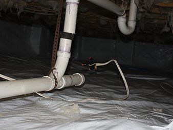 White plumbing pipes and a loose hose in a crawlspace under a house, with plastic sheeting on the floor