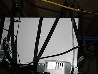 White box device tangled with black cables on a shelf, with a small silver vented unit in front