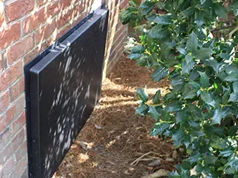 Black wall-mounted utility box beside a brick wall, partly shaded by nearby bushes.
