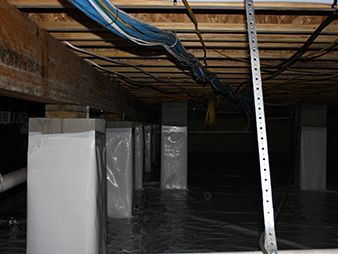 Empty basement with plastic-covered columns, exposed beams, and wires overhead.