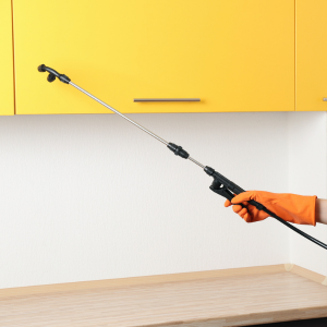 A gloved hand sprays under yellow kitchen cabinets with a long cleaning wand.