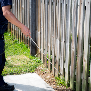 Person using a long-handled tool to trim weeds along a wooden fence in a yard.