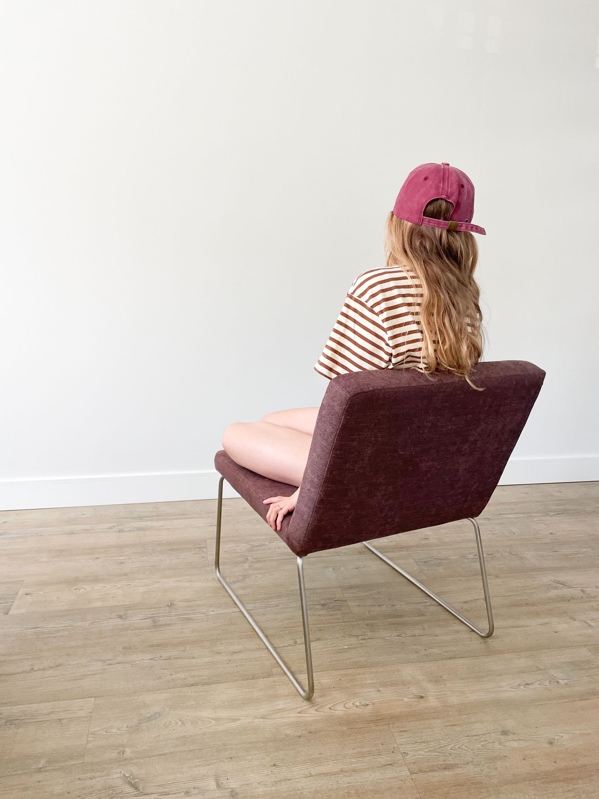 Een persoon zit op een mauve stoel in een witte kamer, van achteren gezien, met een roze pet en een gestreept shirt aan.