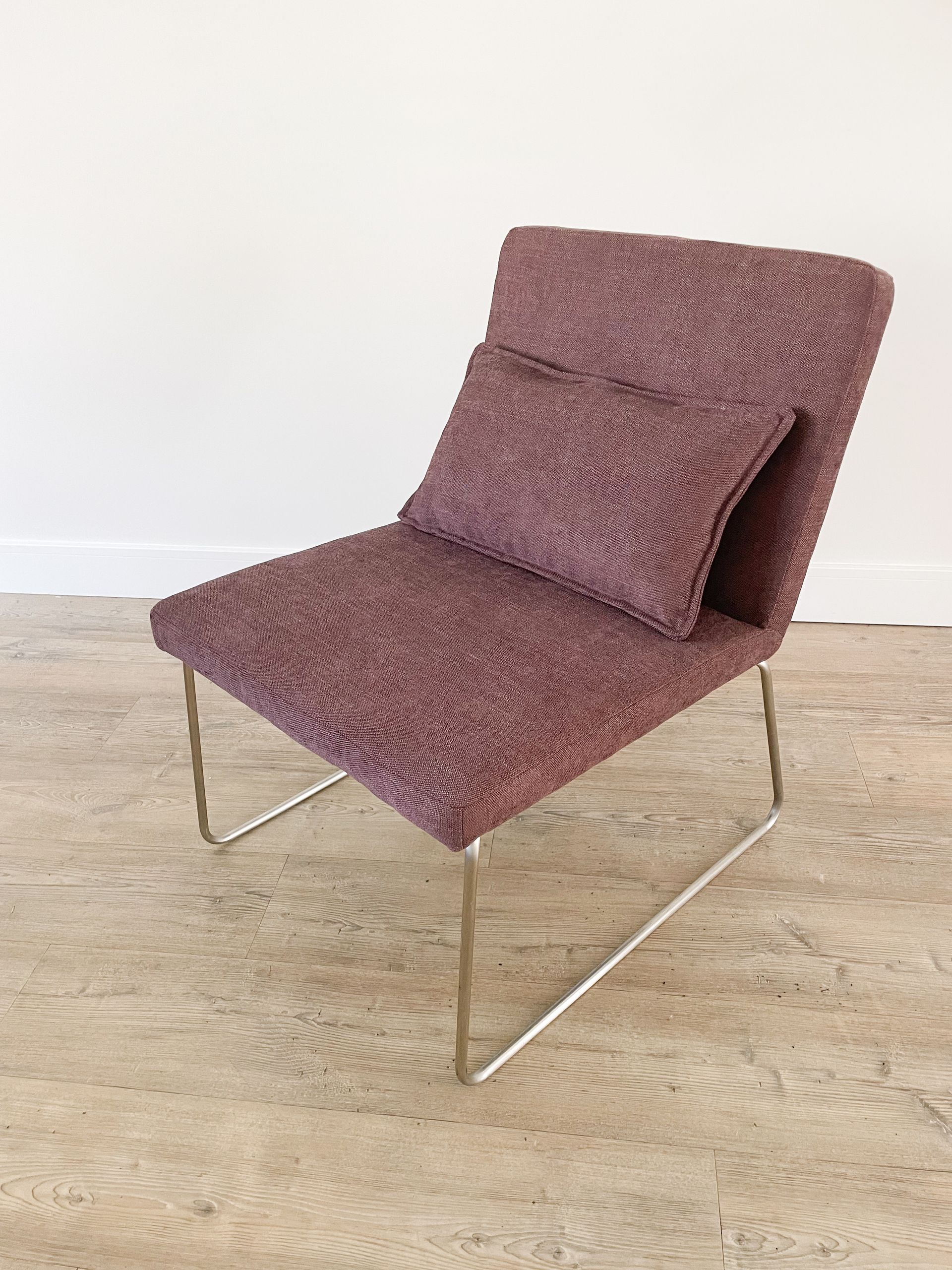 Moderne mauve loungestoel met metalen poten op een lichte houten vloer tegen een witte muur.