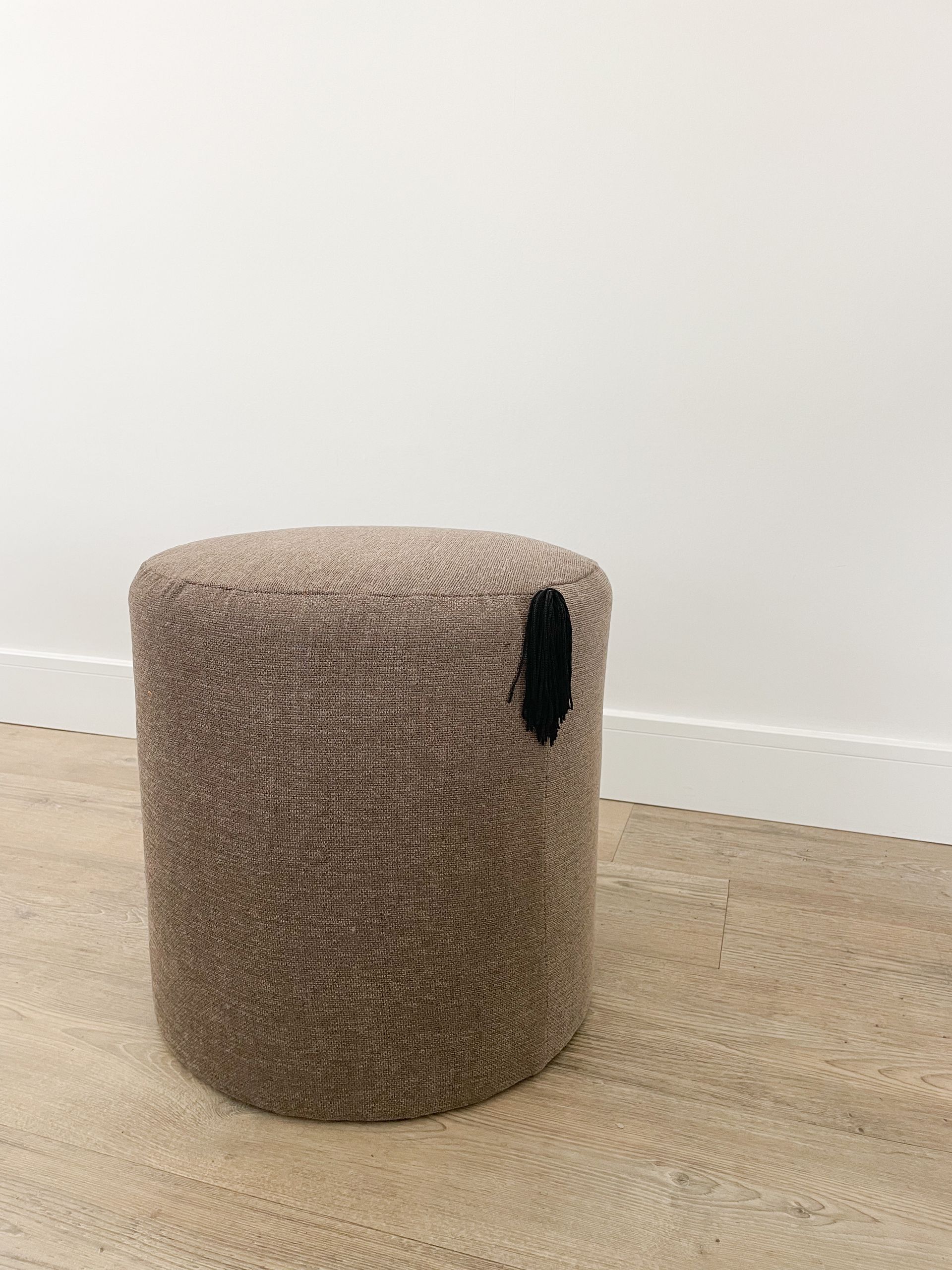 Beige geweven cilindervormige poef op een lichte houten vloer tegen een witte muur.