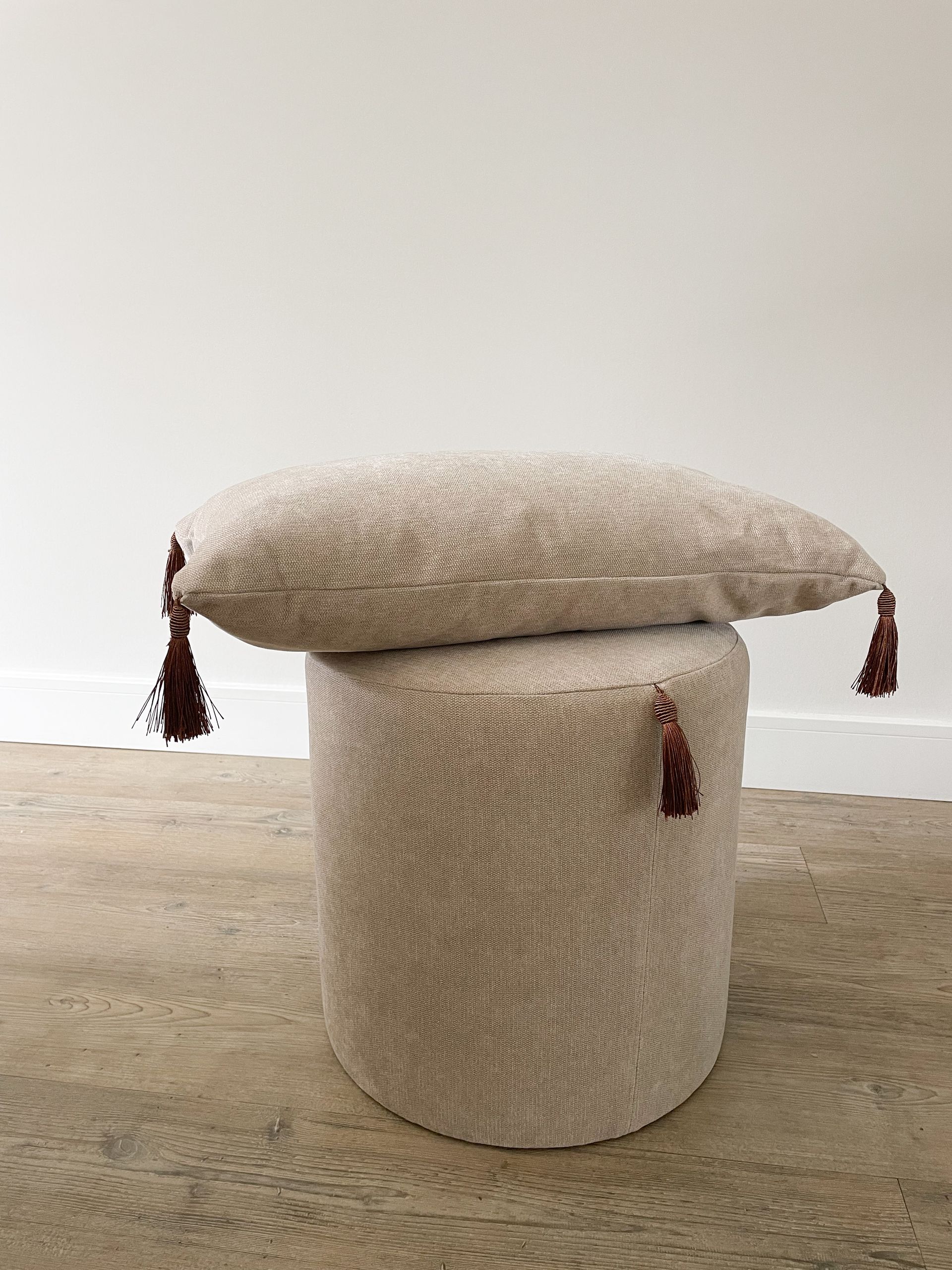 Crèmekleurige, gestructureerde, cilindrische poef op een lichte houten vloer tegen een witte muur, met een klein bruin stoffen labeltje.