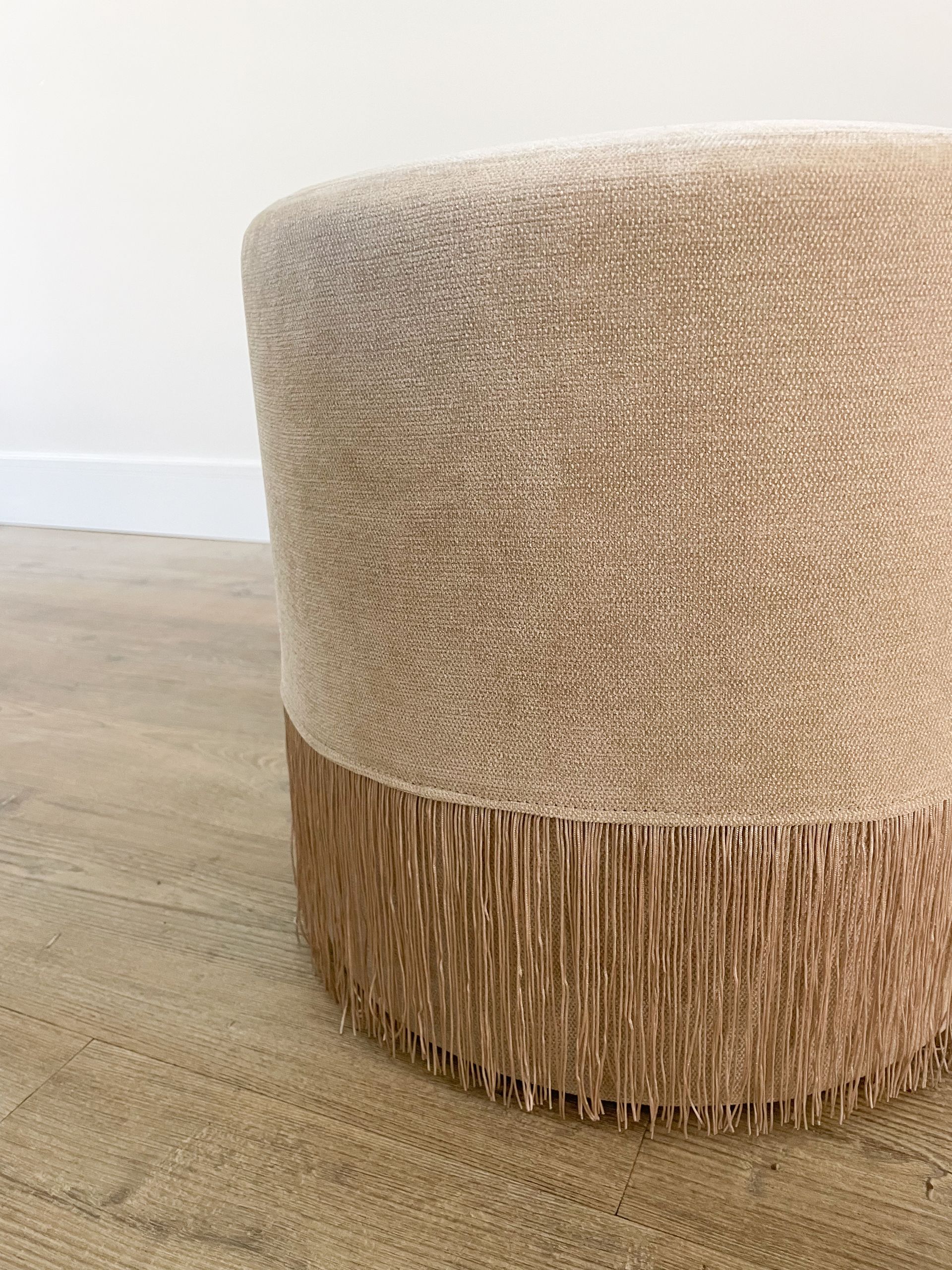 Beige ronde poef met textuur en franjes op een lichte houten vloer tegen een witte muur.
