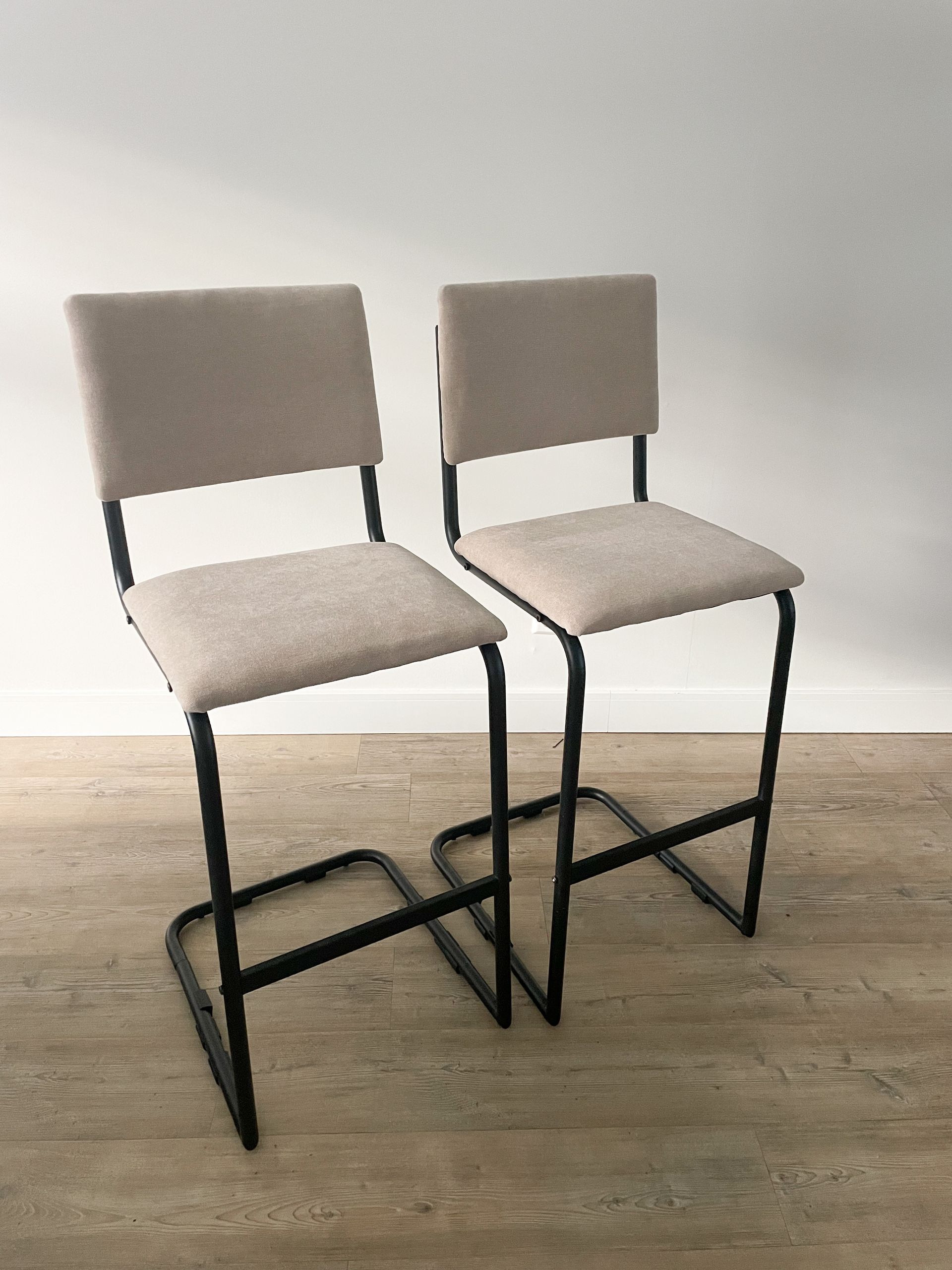 Twee beige gestoffeerde barkrukken met zwarte metalen frames op een lichte houten vloer.