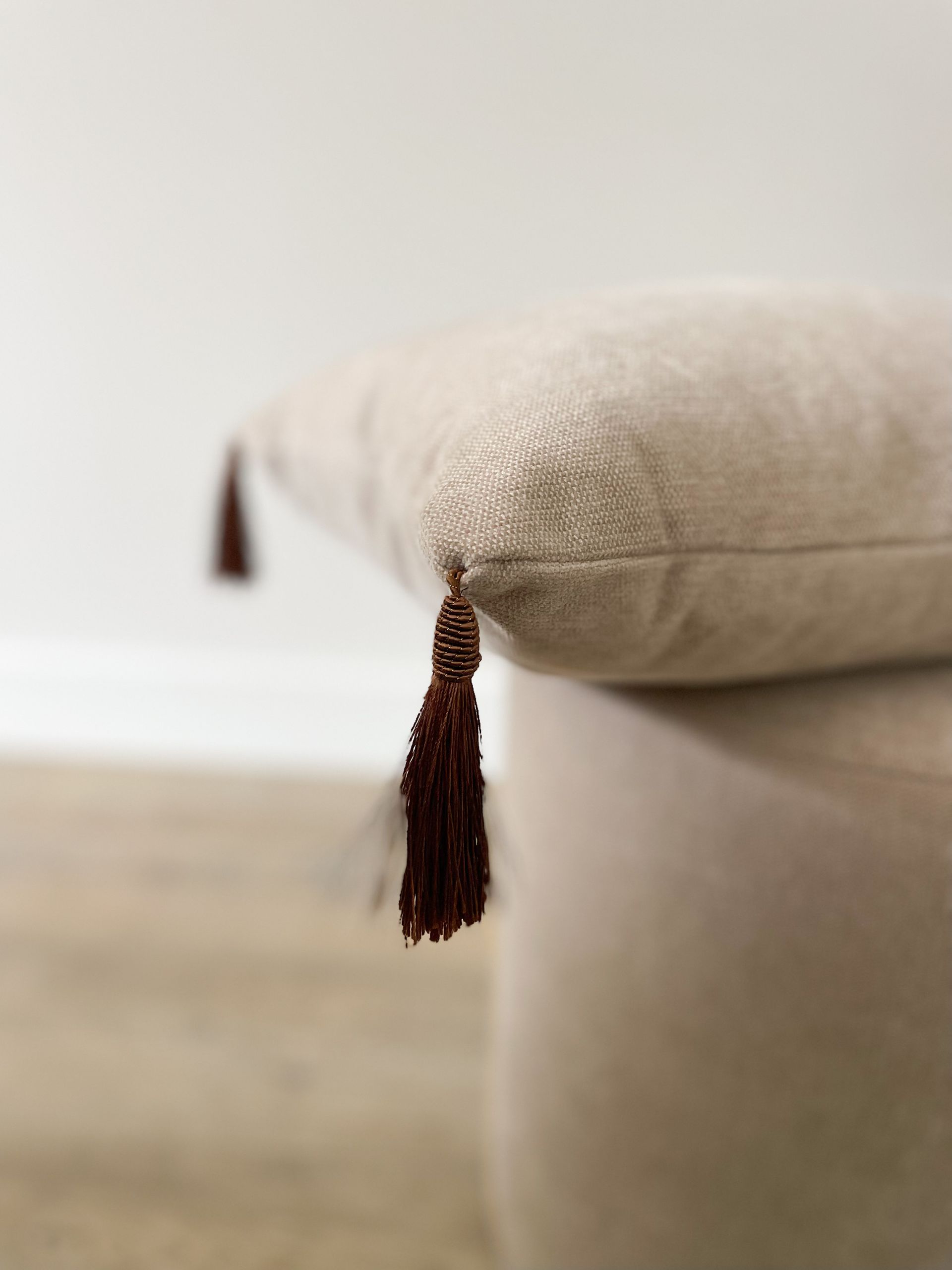 Beige sierkussen met een donkerbruine kwast die boven een lichte houten vloer hangt.