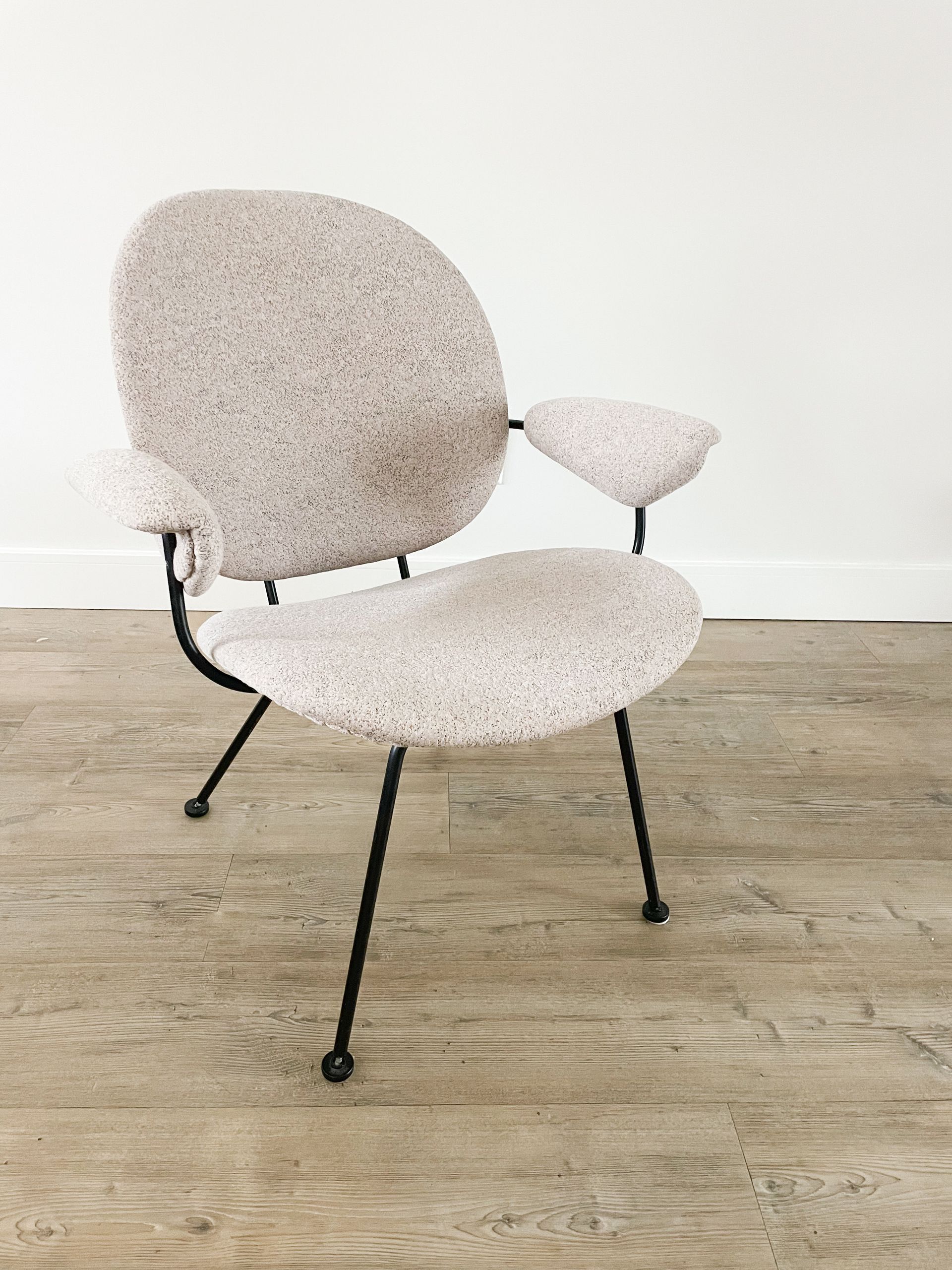 Beige stoel met structuur, zwarte poten en een klein bijzettafeltje op een lichte houten vloer tegen een witte muur.