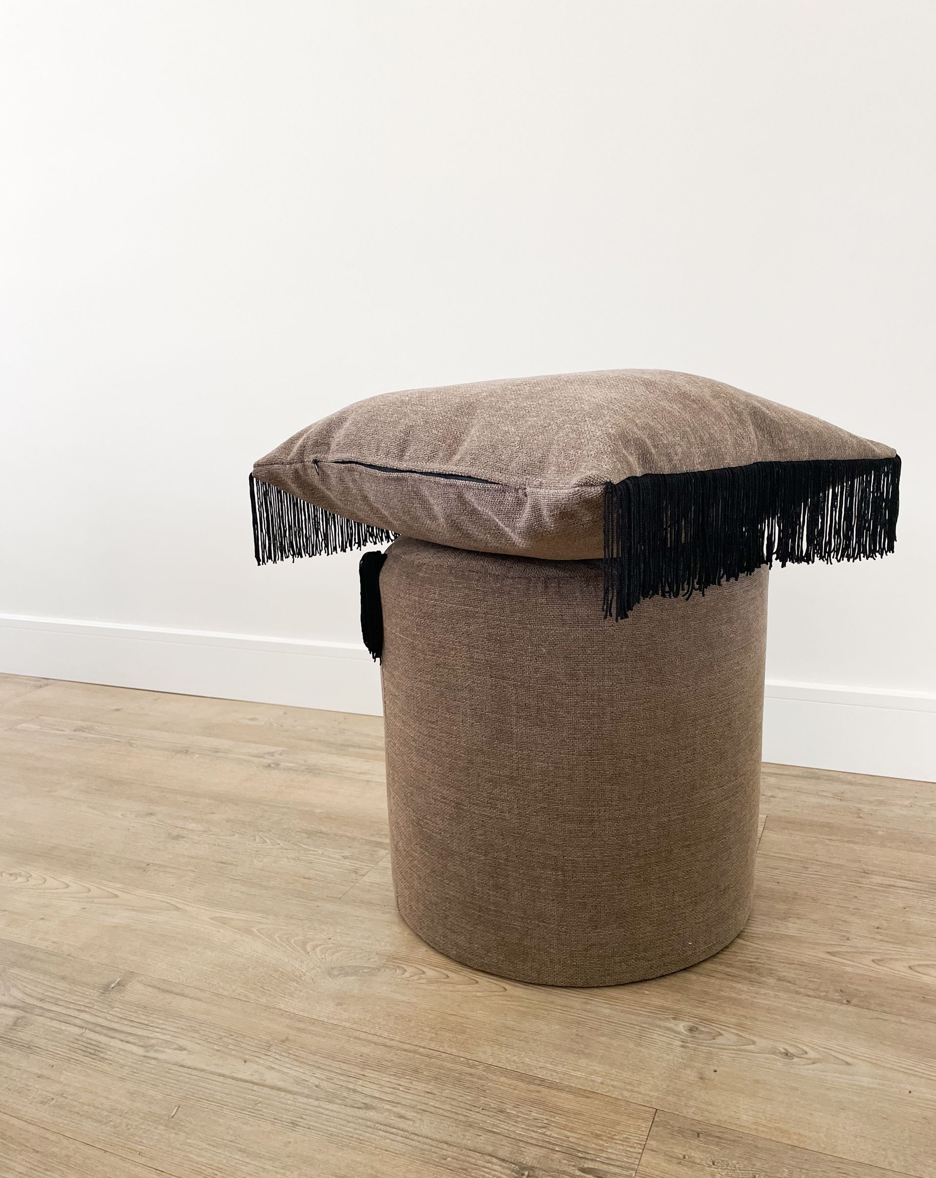 Bruine, gestructureerde kruk met een gewatteerde zitting en zwarte franjes, op een lichte houten vloer tegen een witte muur.