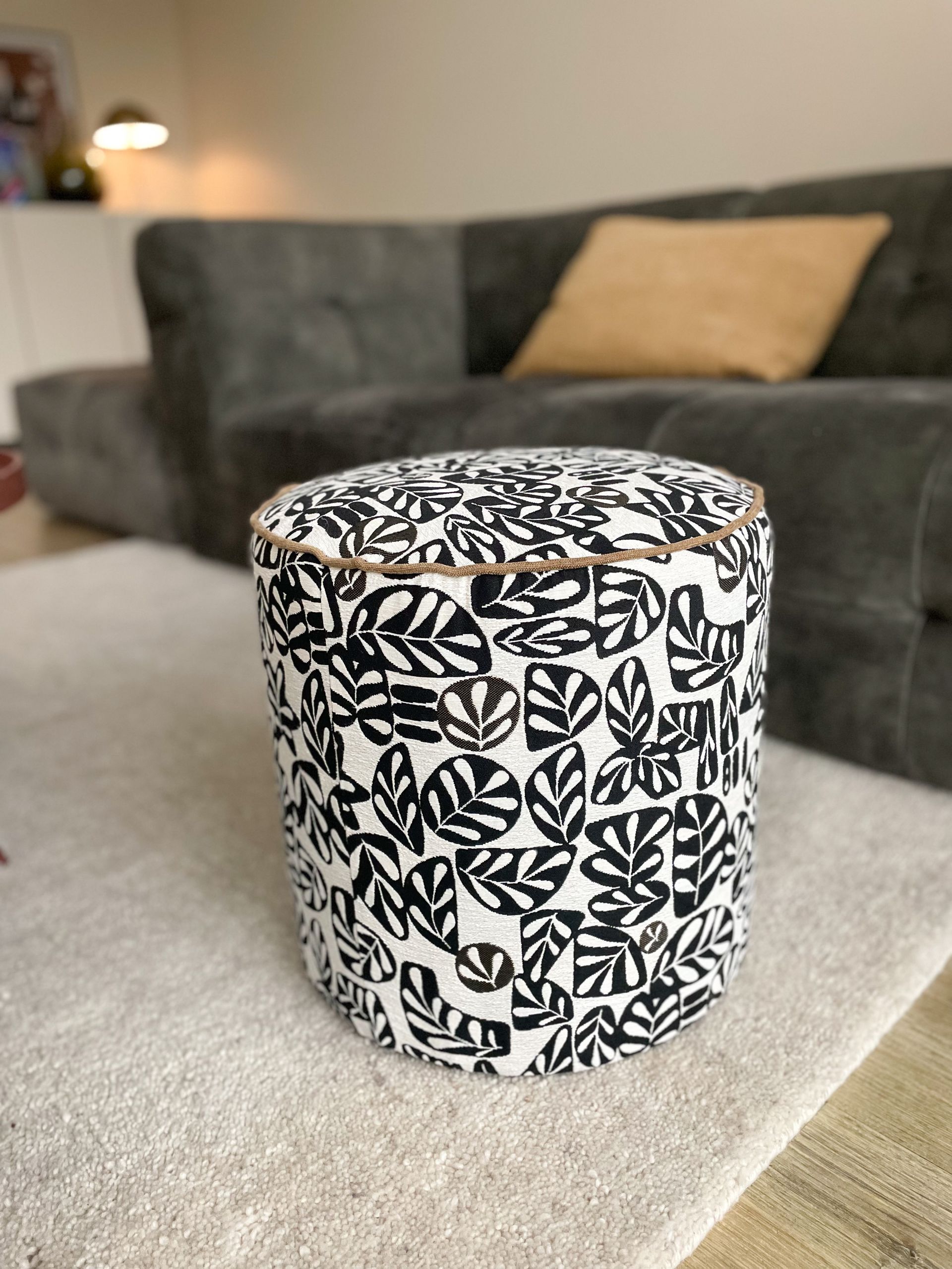 Een cilindervormige poef met zwart-wit patroon op een grijze bank in een woonkamer.
