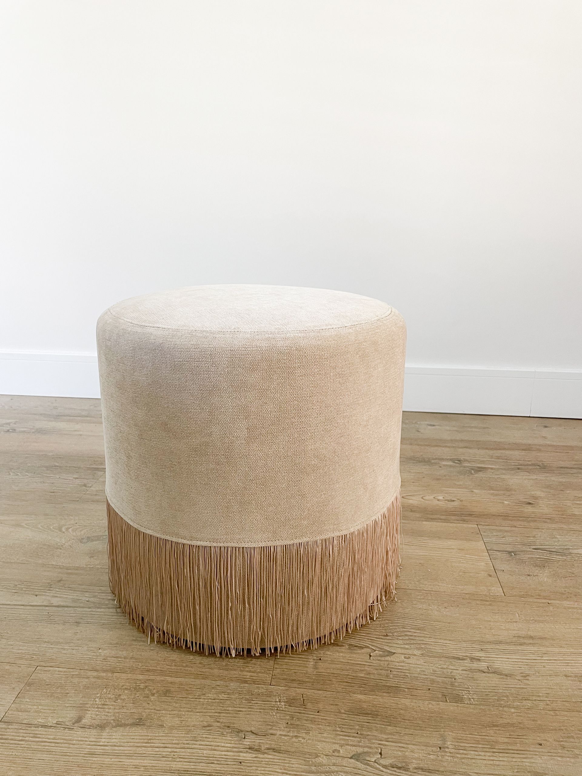 Ronde, beige en lichtbruine poef met textuur op een betonnen vloer tegen een witte muur.