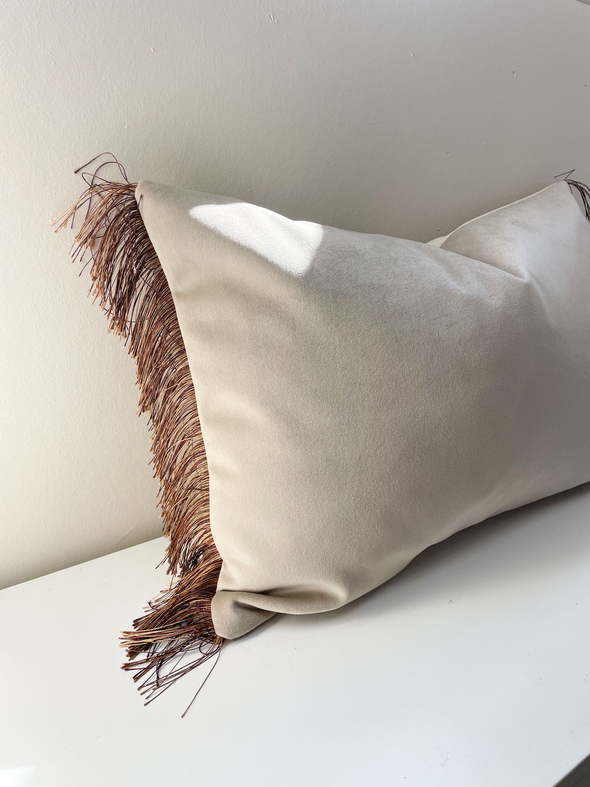 Beige kussen met bruine geweven franjes op een witte bank tegen een lichte muur.