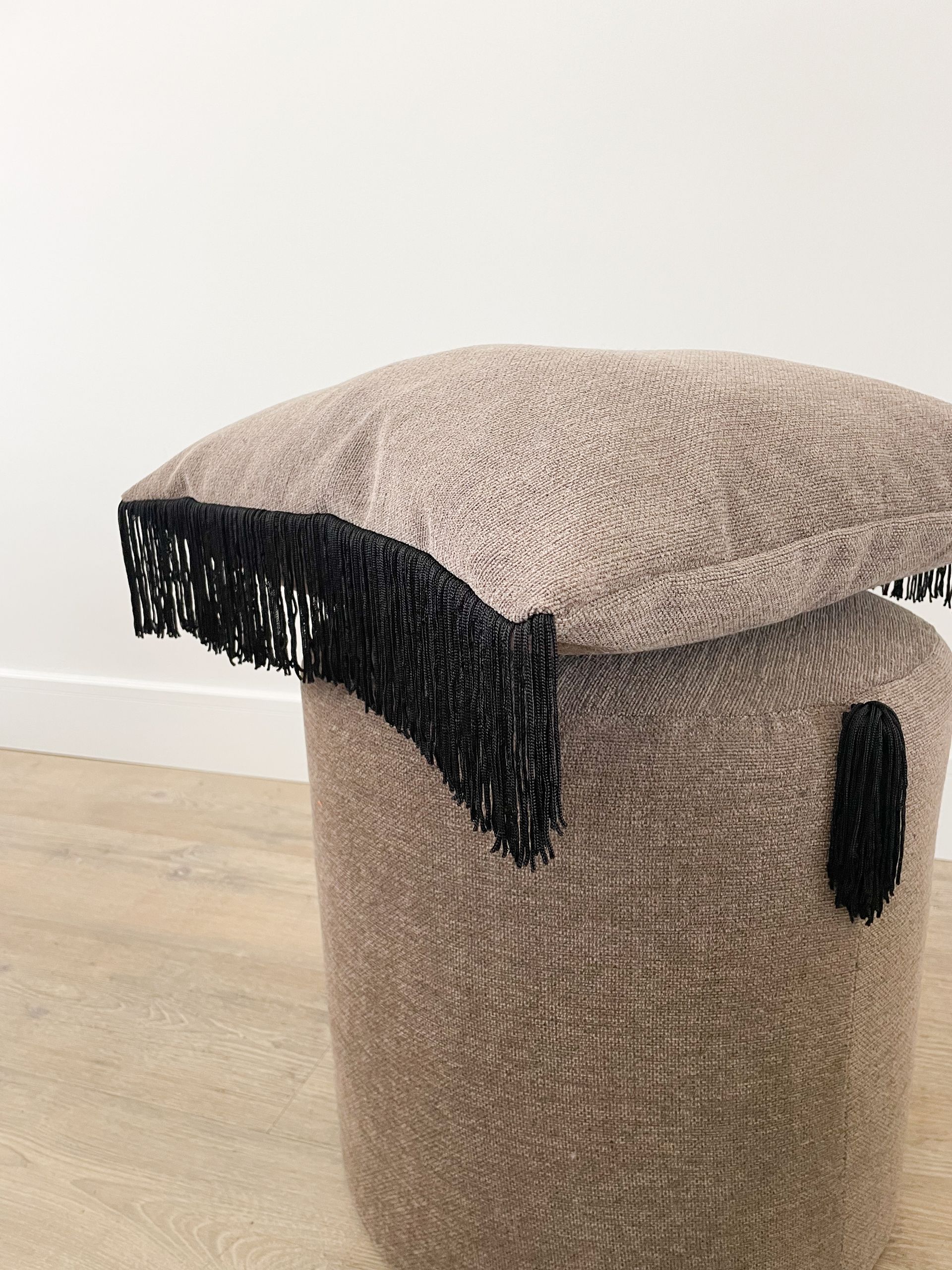 Een beige poef met structuur, een beige kussen en zwarte franjes op een lichte houten vloer.