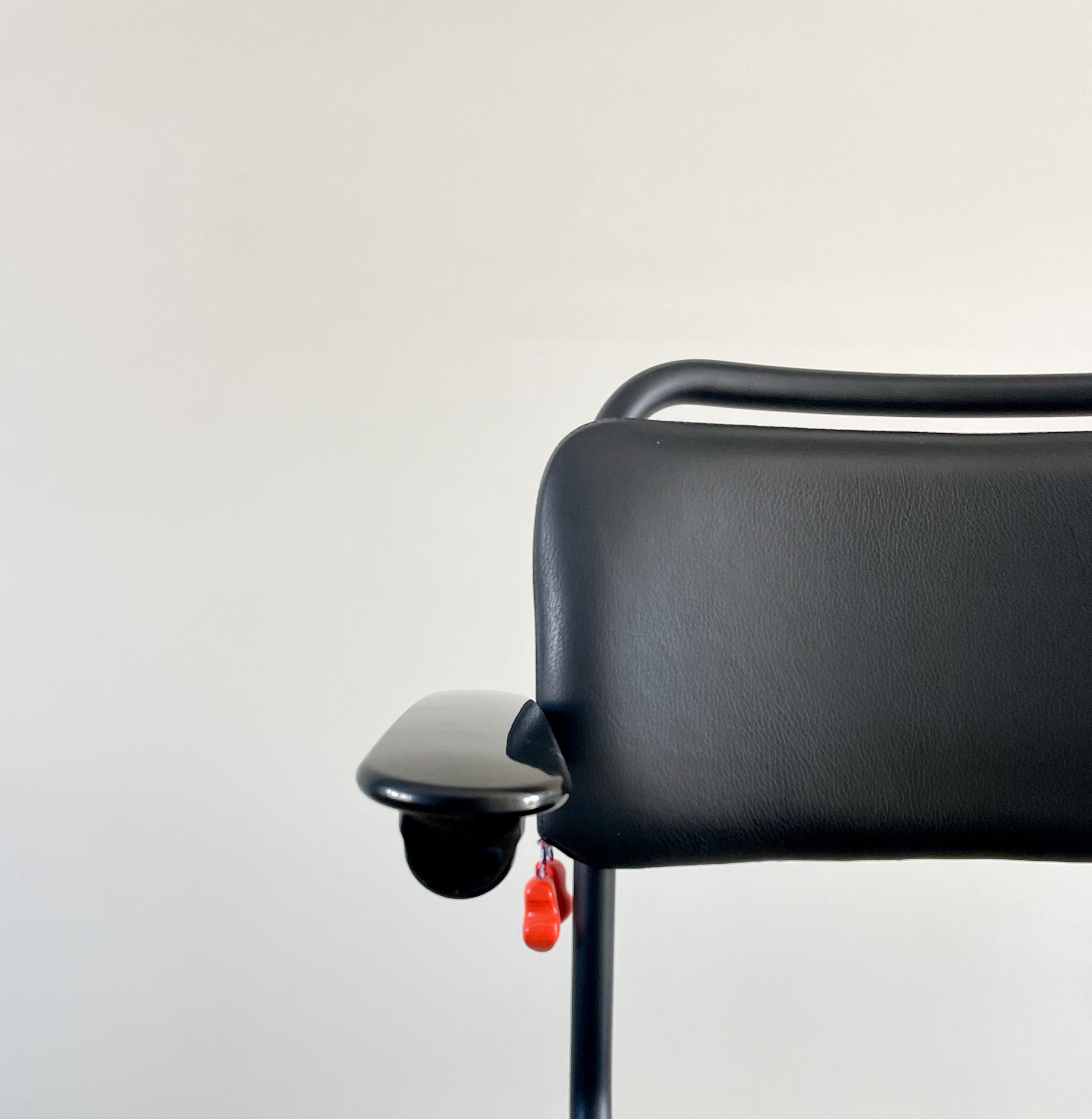 Zwarte armleuning van een bureaustoel met een klein rood labeltje, tegen een effen muur.