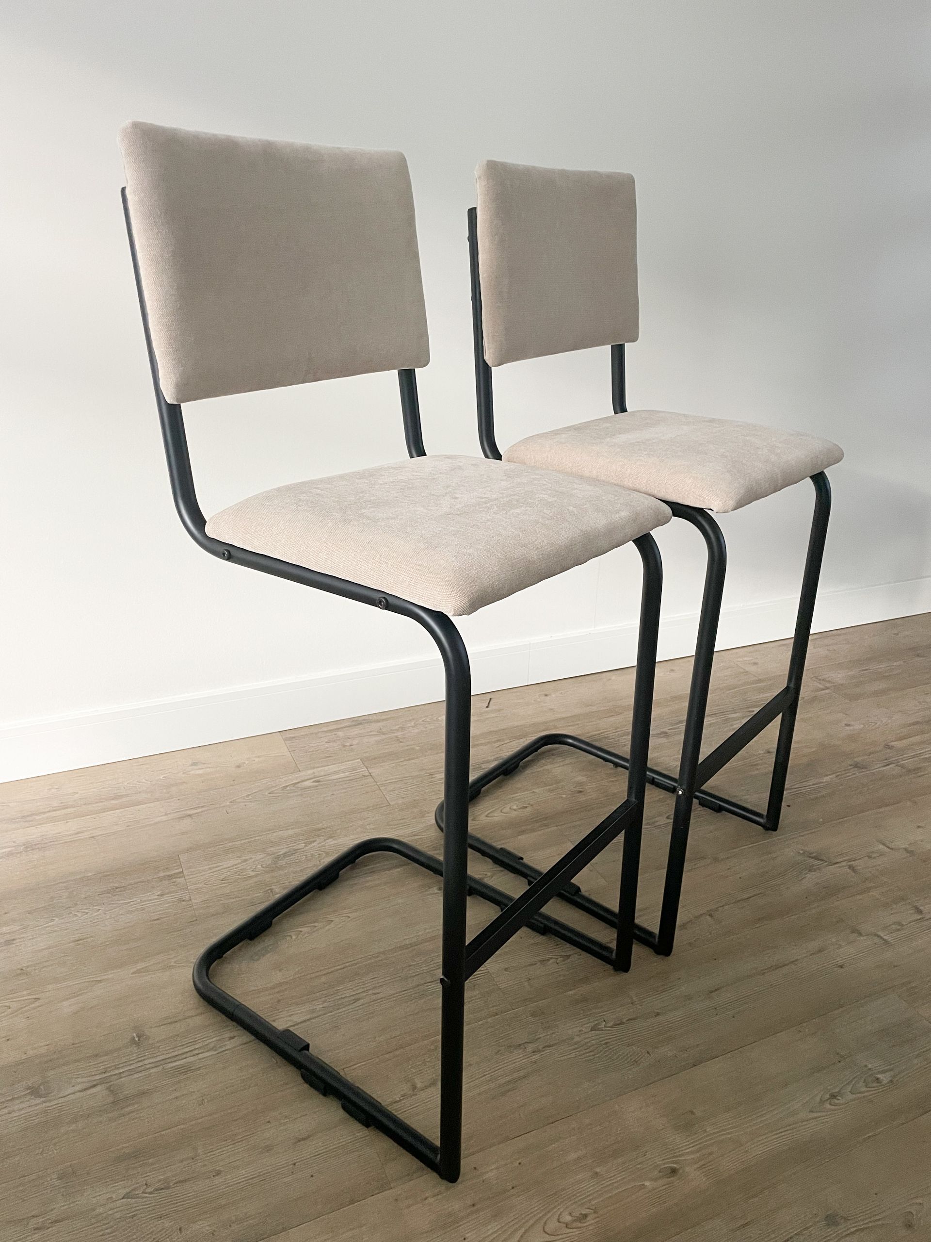 Twee beige gestoffeerde barkrukken met zwarte metalen frames op een houten vloer tegen een witte muur.